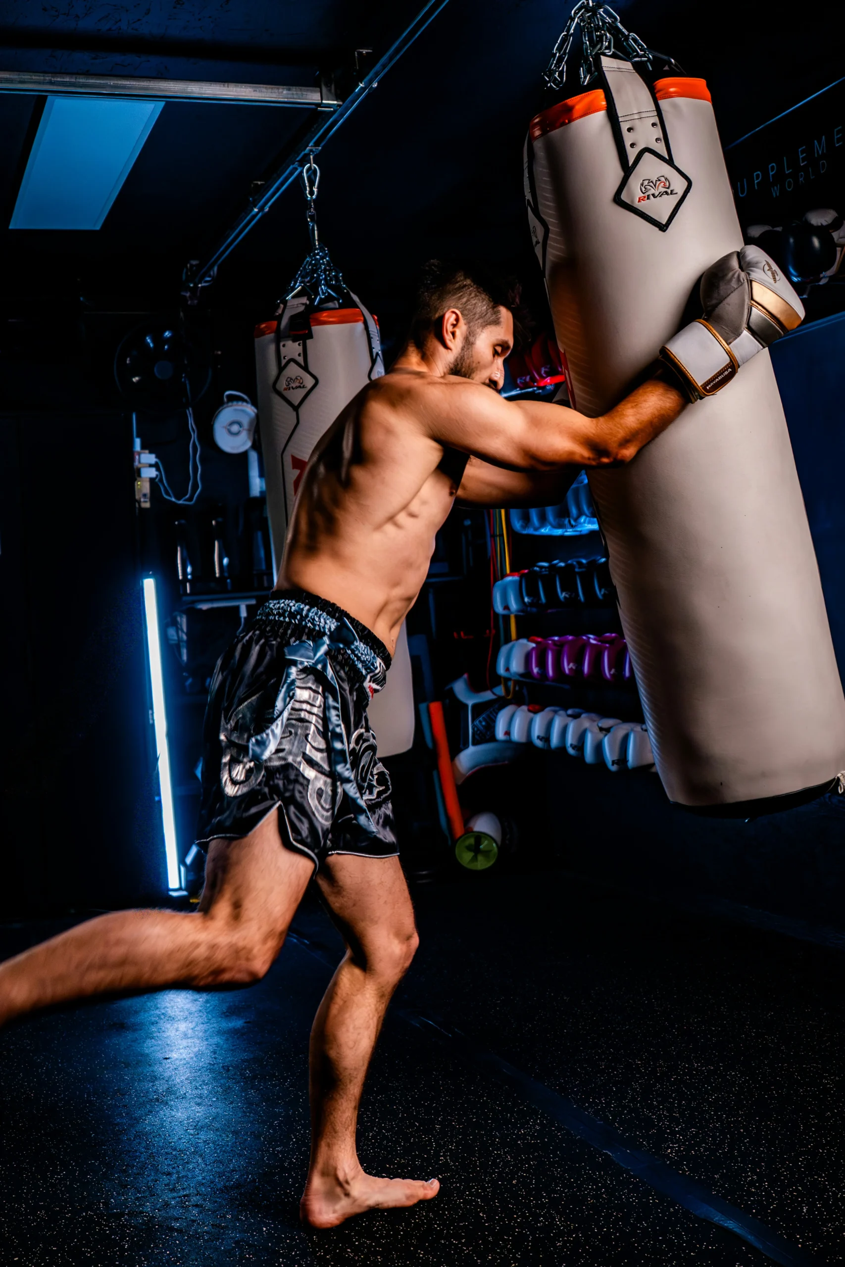 Muay Thai Shorts Manufacturer showcasing a male fighter wearing black satin Muay Thai shorts while training on a heavy bag in a boxing gym.