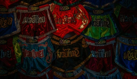 Custom Muay Thai Shorts in colourful satin designs with embroidered Thai lettering, decorative trims, and traditional wide waistbands.