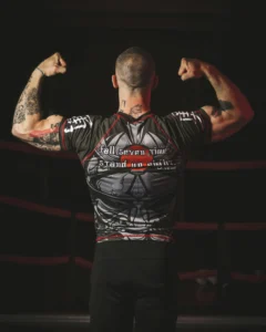 Male athlete wearing a black short-sleeve rash guard and flexing inside a combat sports ring.